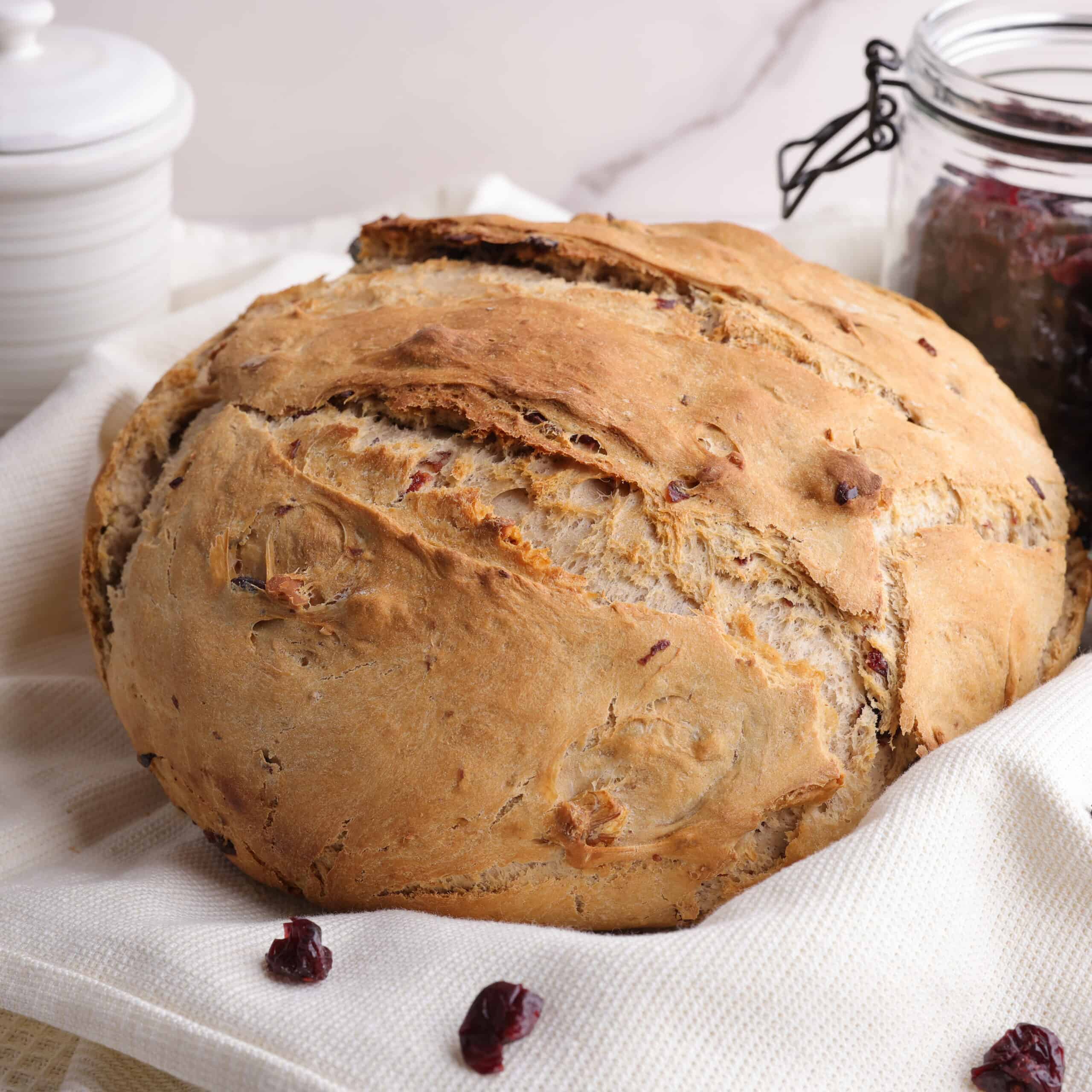 A whole cooked loaf of cranberry walnut bread decoratively displayed on a tea towel with a jar of honey and craisins in the background to demonstrate some of it's ingredients.