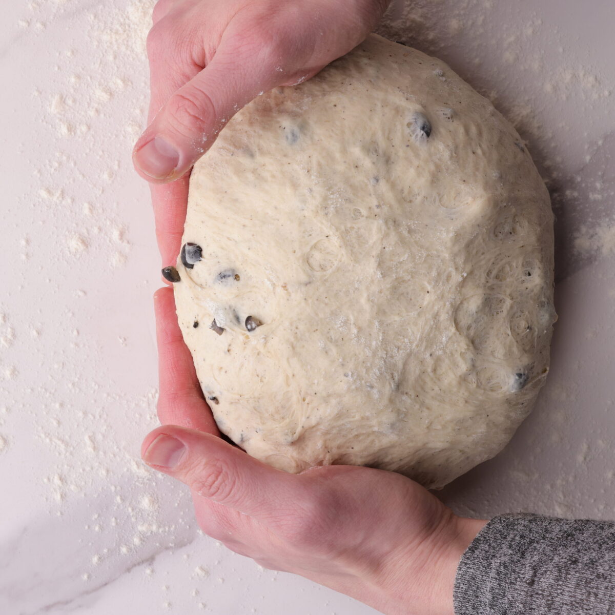 A ball of bread dough on a counter sprinkled with flour being pulled inward by two hands which are cupping the top of the ball of dough to shape the bread.