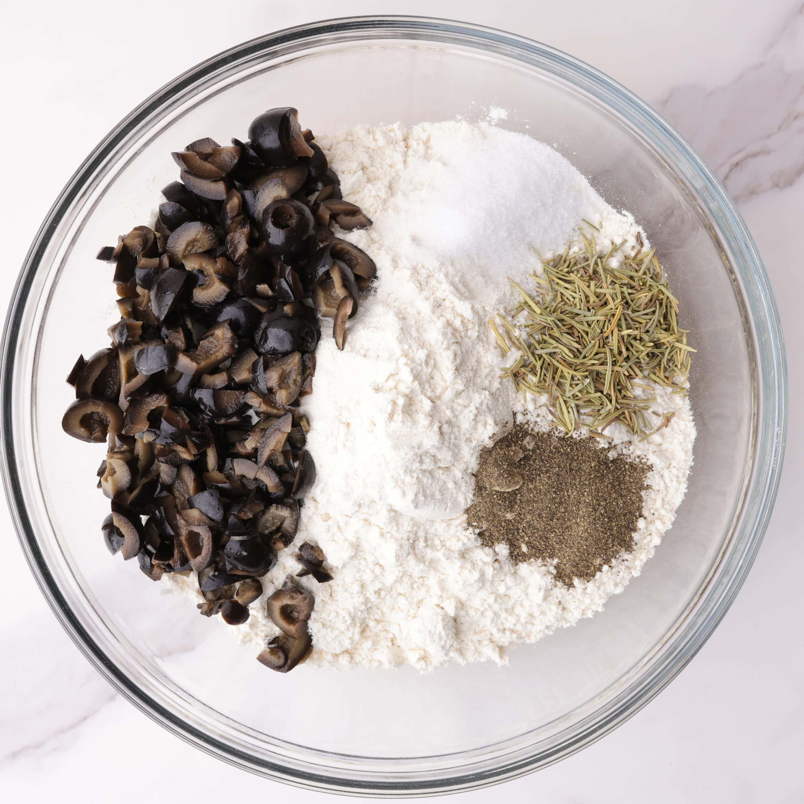 A mixing bowl with all dry ingredients - flour, salt, pepper, olives, and dried rosemary - in it.
