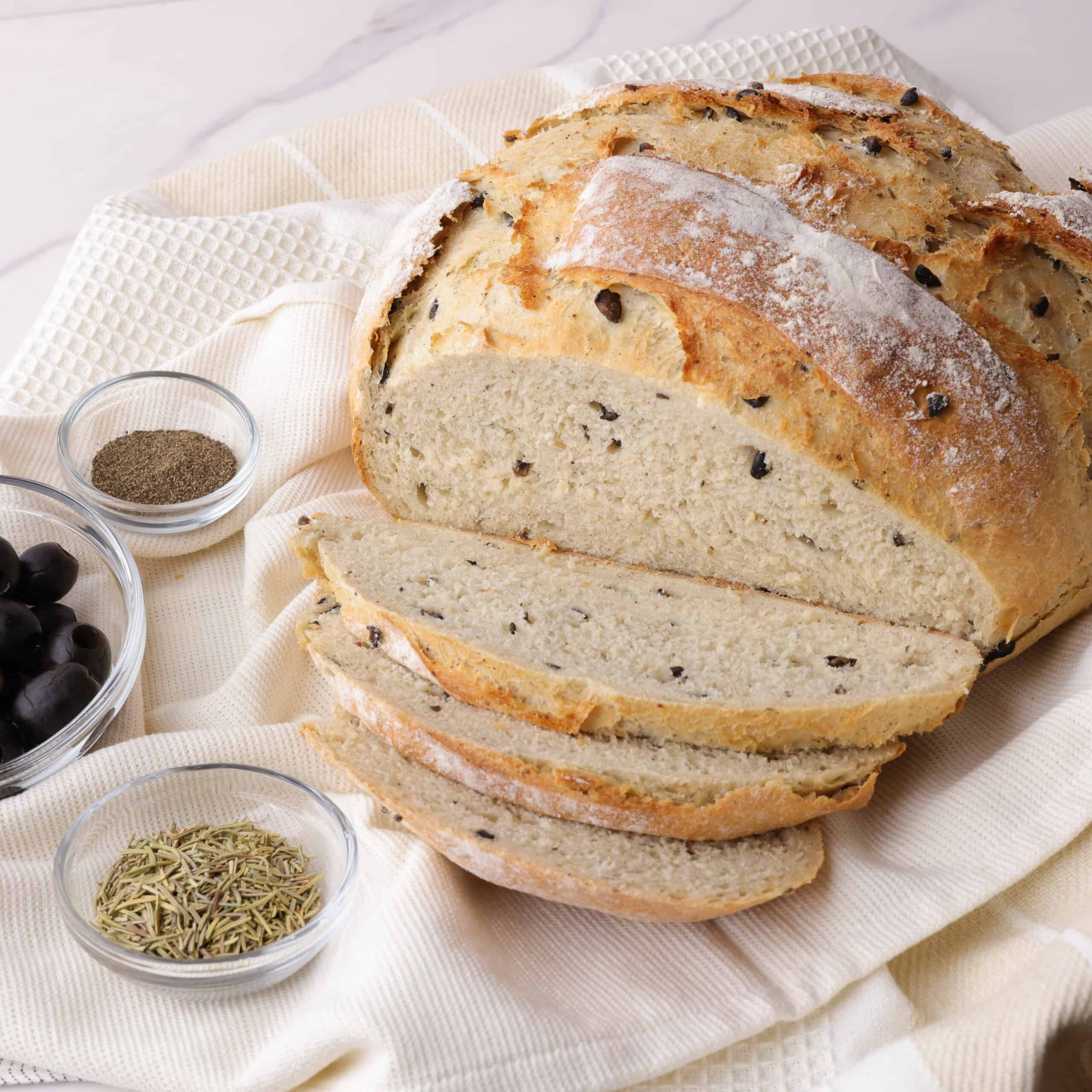 A decorative display of the Rosemary Garlic bread, partially sliced on a tea towel with bowls of rosemary, olives and pepper on the side to demonstrate some of the ingredients. 