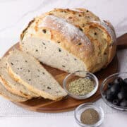 A decorative display of a baked loaf of the Rosemary Garlic Bread partially sliced on a charcuterie board with bowls of dried rosemary, pepper and olives to demonstrate some of the tasty ingredients.