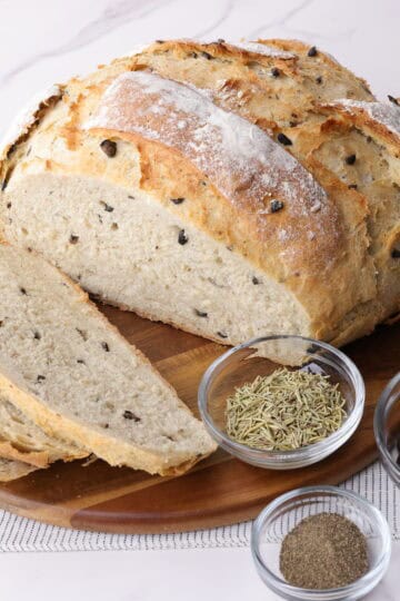 A decorative display of a baked loaf of the Rosemary Garlic Bread partially sliced on a charcuterie board with bowls of dried rosemary, pepper and olives to demonstrate some of the tasty ingredients.