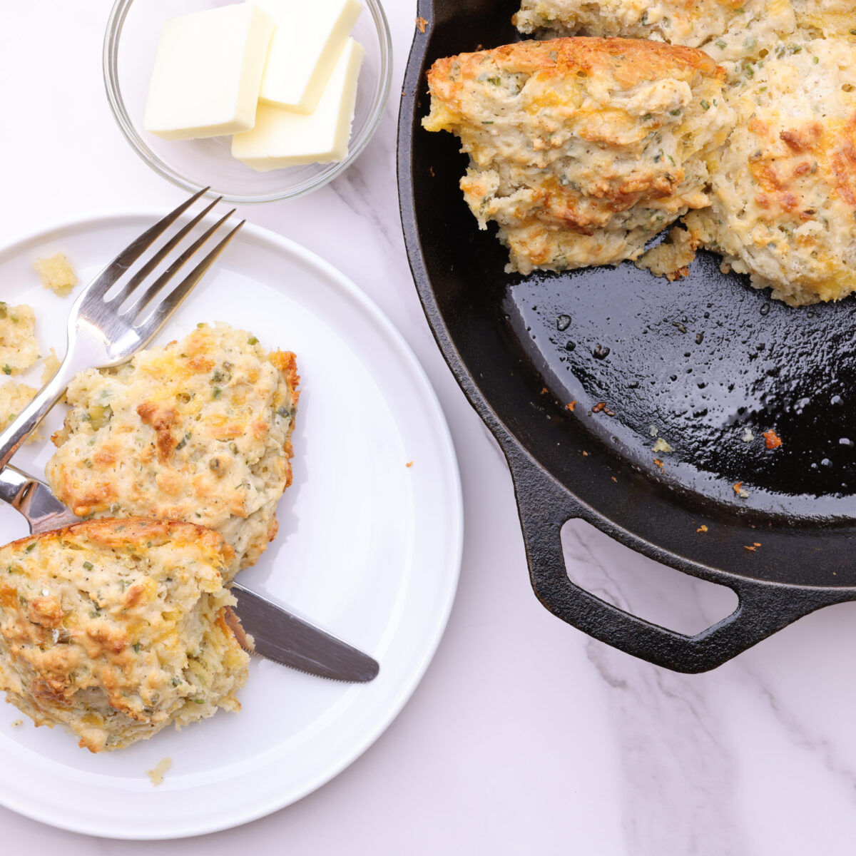 A decorative display of the cooked cheddar cheese chive biscuits with two on a plate with a fork and knife and the skillet with the remaining biscuits beside it with a bowl of butter in the corner.