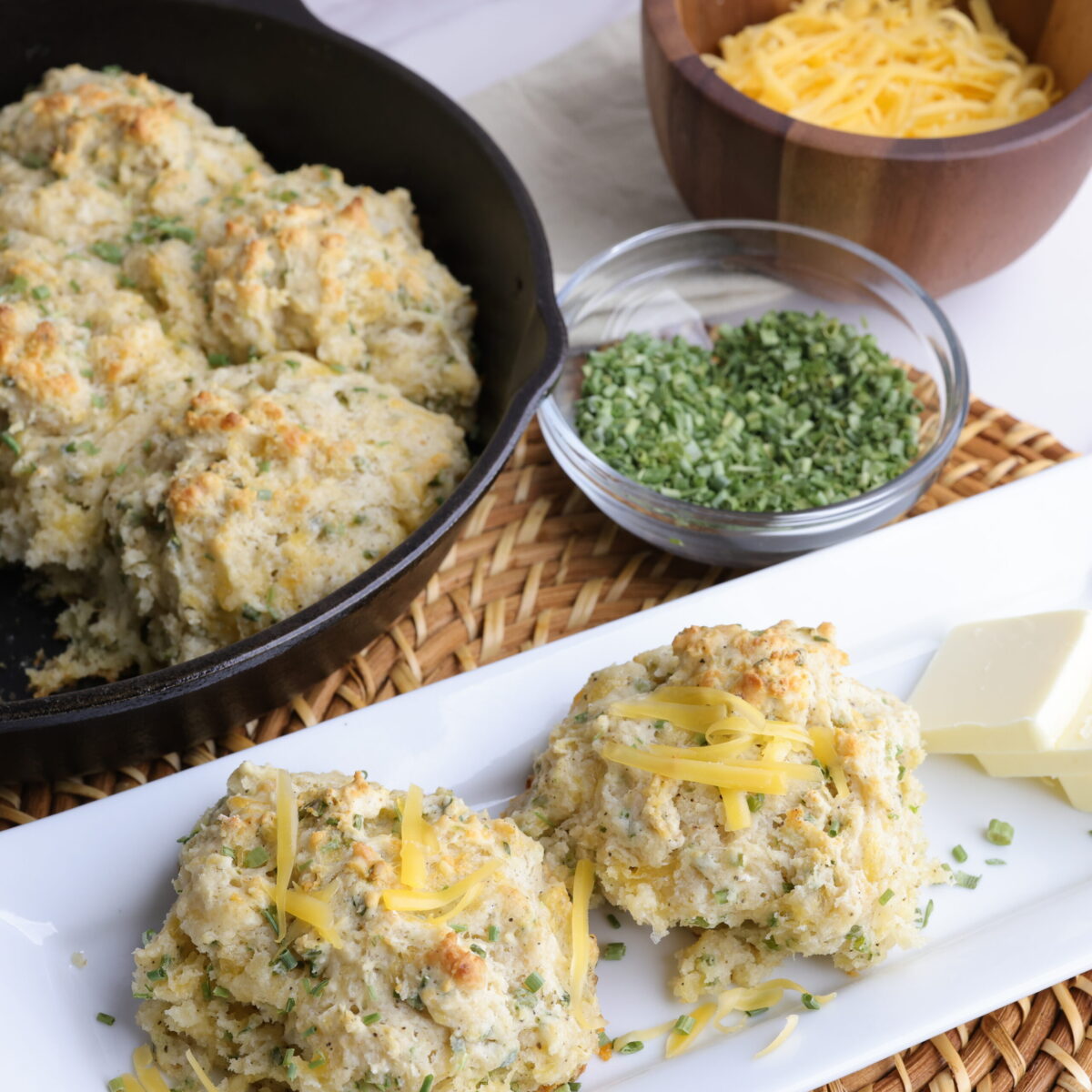 A decorative display of the Cheddar Cheese Chive biscuits with some in the skillet they were cooked in, some displayed on a plate sprinkled with cheese and side of butter and a bowl of chives and cheddar cheese to demonstrate some of the fillings.