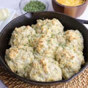 A skillet with baked cheddar cheese chive biscuits sitting on a basket weave place mat with sliced butter, dried chives and cheddar cheese in the background.