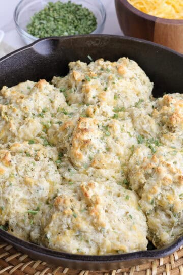 A skillet with baked cheddar cheese chive biscuits sitting on a basket weave place mat with sliced butter, dried chives and cheddar cheese in the background.