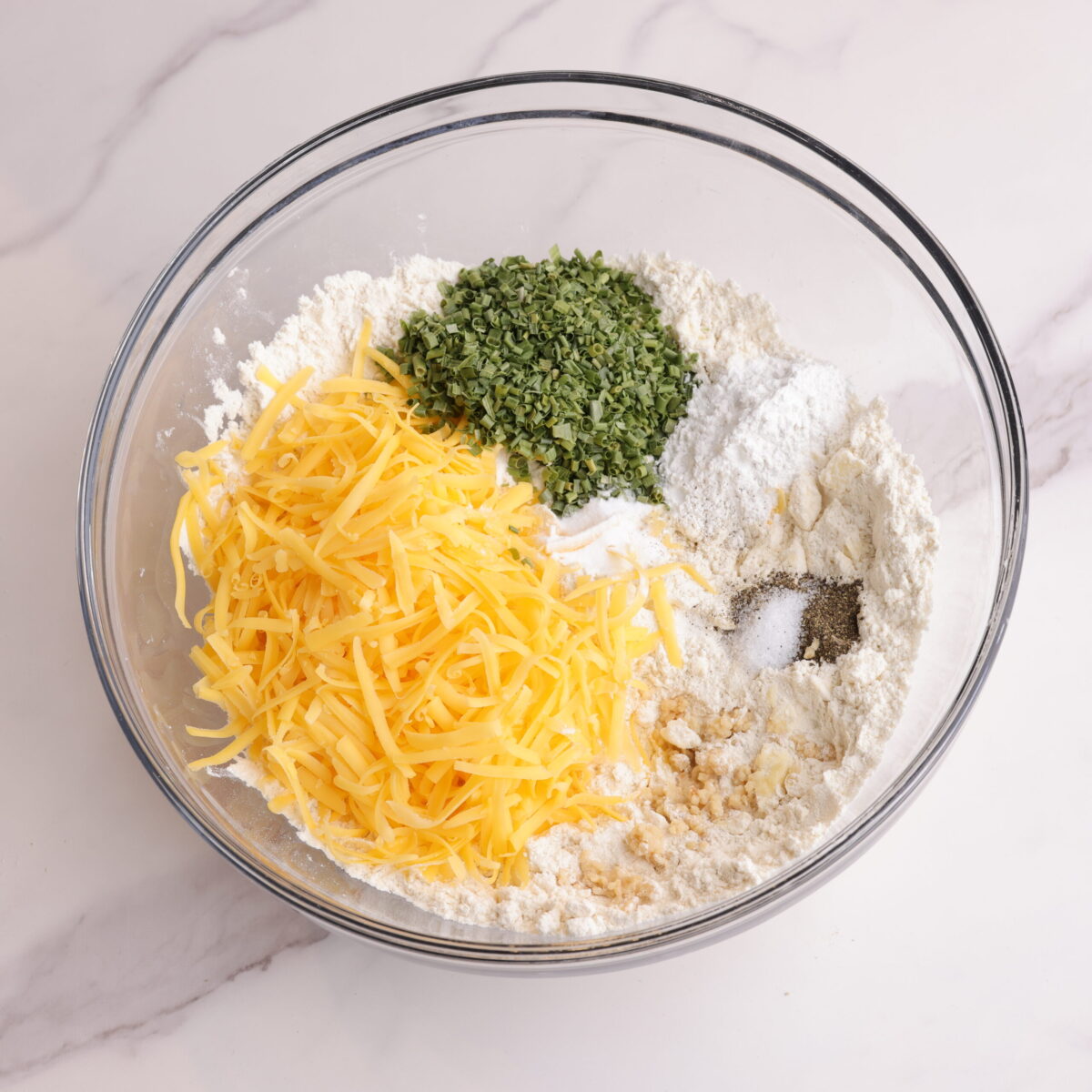 The bowl with the flour and butter cut in now topped with the shredded cheddar cheese, dried chives, baking powder, baking soda, garlic, salt and pepper.