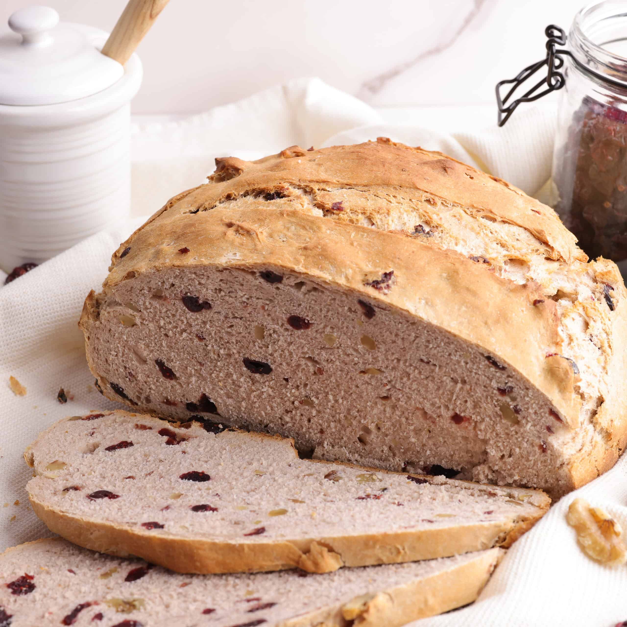 Loaf of cranberry walnut bread, sliced and displayed on a tea towel with a jar of honey and craisins in the background to demonstrate some of it's fillings.