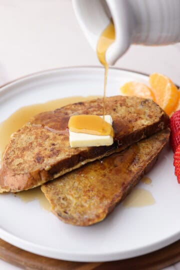 Delicious Cranberry Walnut French toast plated with a cube of butter on top and sweet maple syrup being poured over the top. Two strawberries and orange slices are decoratively placed along the side of the plate.