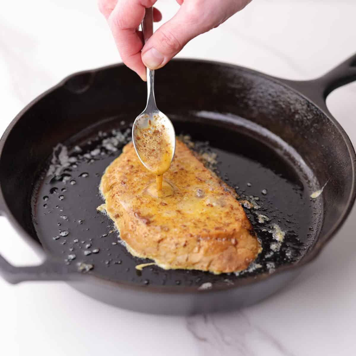 Cranberry walnut french toast cooking in a cast iron skillet with the egg and spice mixture being poured on to coat the second side of the bread before flipping.