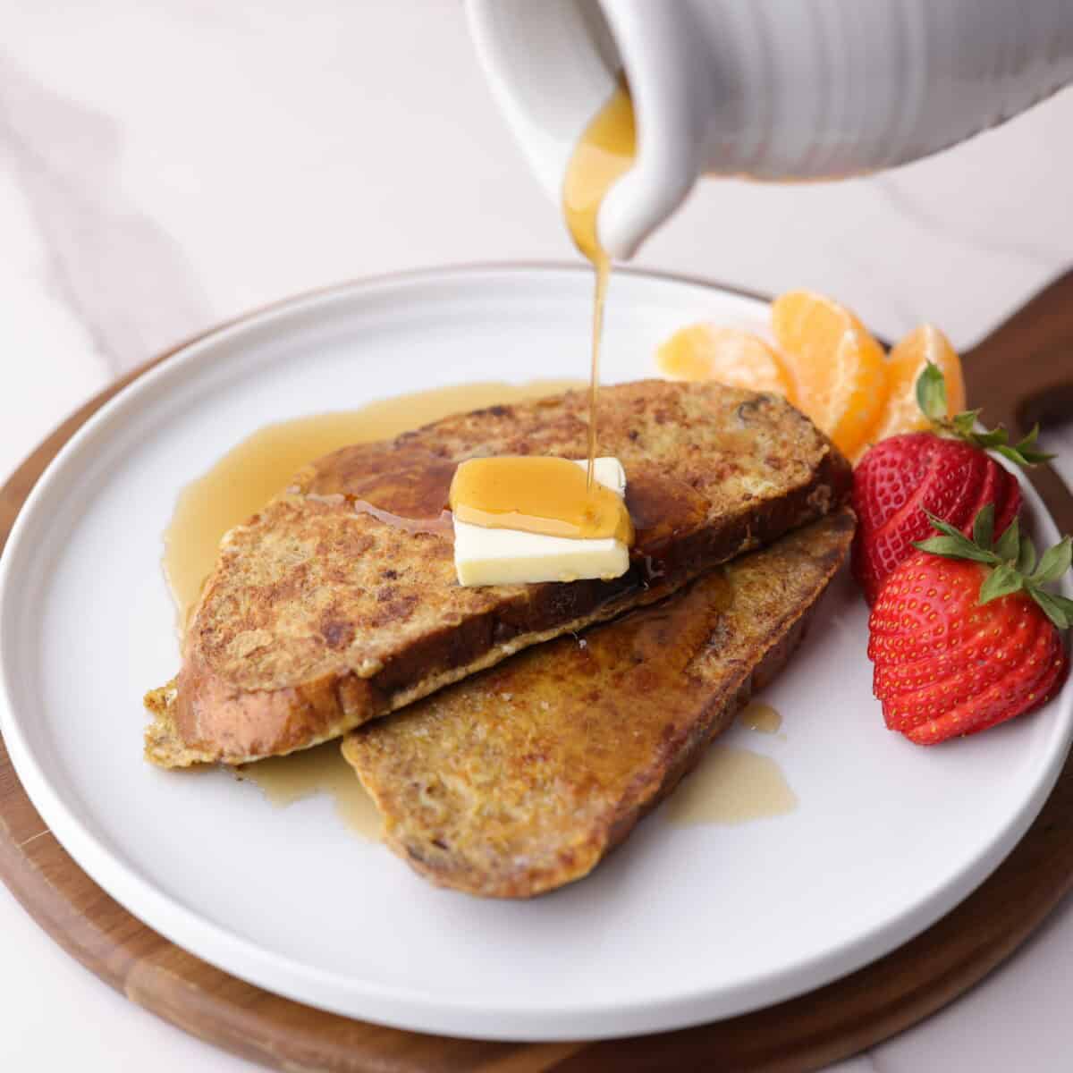Delicious Cranberry Walnut French toast plated with a cube of butter on top and sweet maple syrup being poured over the top. Two strawberries and orange slices are decoratively placed along the side of the plate.