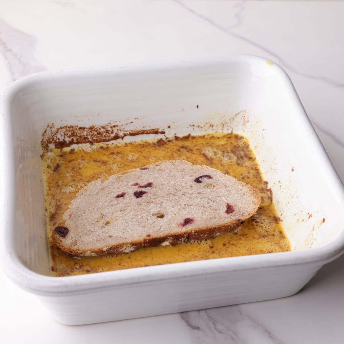 Cranberry walnut bread being coated in the egg and spice mixture in a dish for making French toast.
