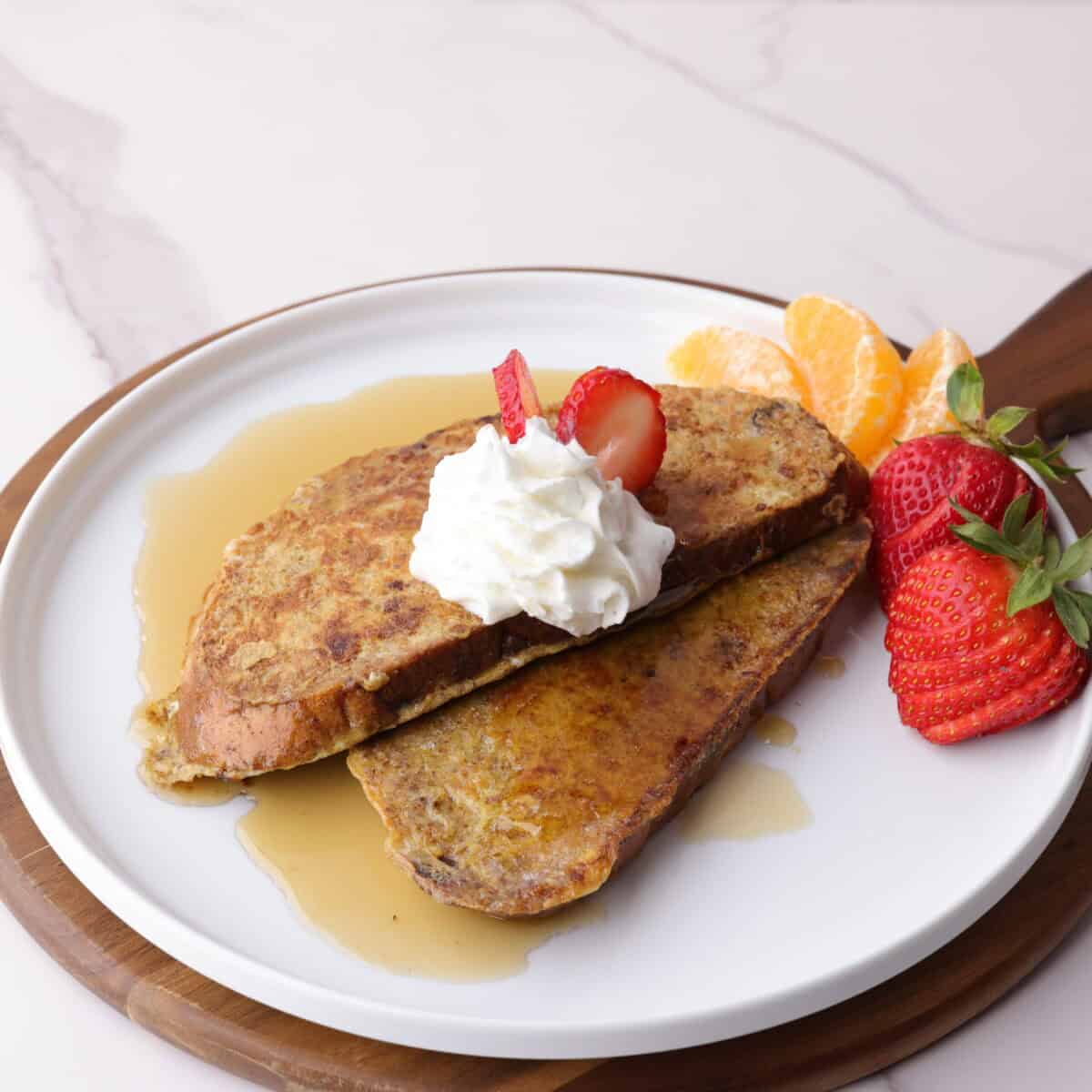 Plated cranberry walnut french toast with whipped cream, drizzled in sweet maple syrup and arranged with strawberries and orange sliced on the side.