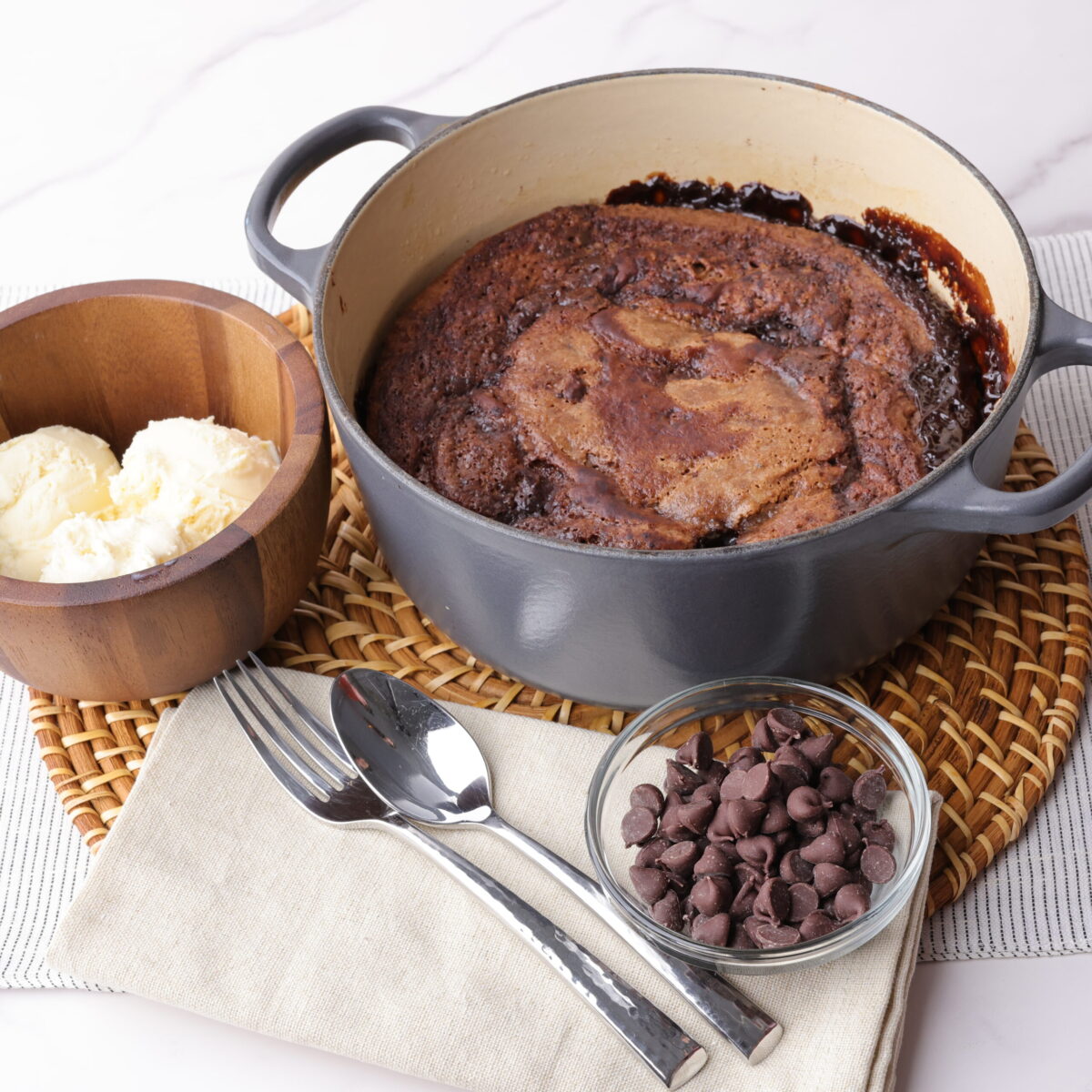 Deliciously chocolatey Fudge Pudding in an enameled cast iron pot decoratively laid out with bowl of ice cream and bowl of chocolate chips on a place mat.