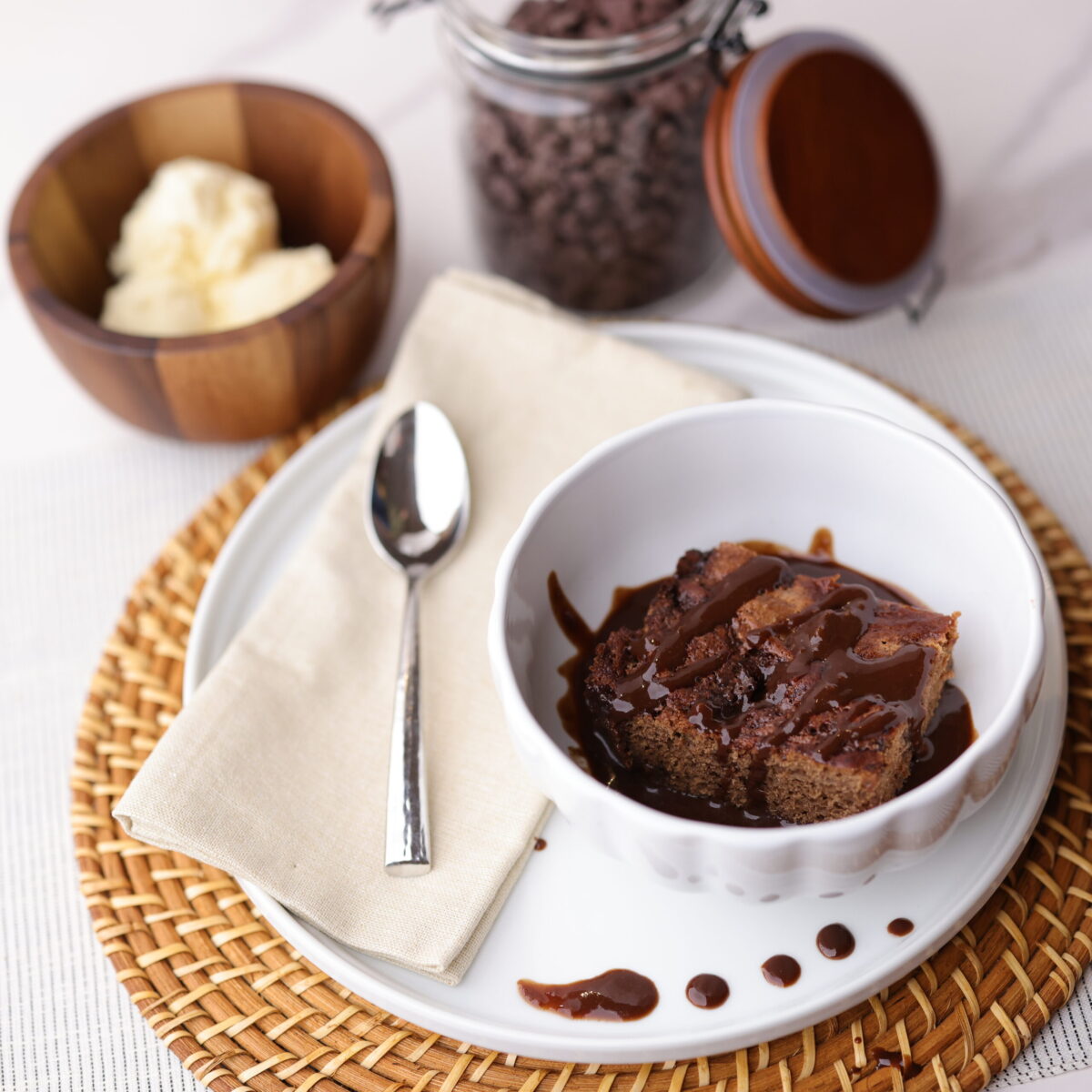 A piece of the baked Fudge pudding in bowl, drizzled with fudge sauce placed decoratively on a plate with a napkin and spoon. A bowl of ice cream in a wooden bowl with chocolate chips in a separate jar are on the side.