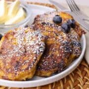 Perfectly browned pumpkin cottage cheese pancakes on a plate, sprinkled with powdered sugar, topped with blueberries and bananas and a small dish of cubed butter on the side.