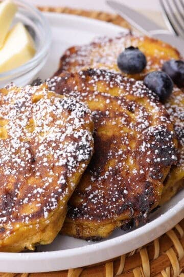 Perfectly browned pumpkin cottage cheese pancakes on a plate, sprinkled with powdered sugar, topped with blueberries and bananas and a small dish of cubed butter on the side.