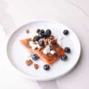 Perfectly baked salmon topped with goat cheese, pecans, and blueberries on white plate.