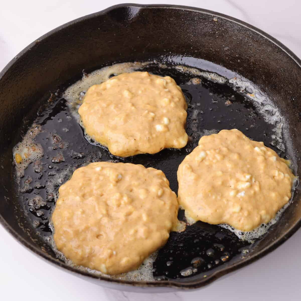 Pumpkin cottage cheese pancake batter spooned into a hot cast iron skillet forming three small pancakes.