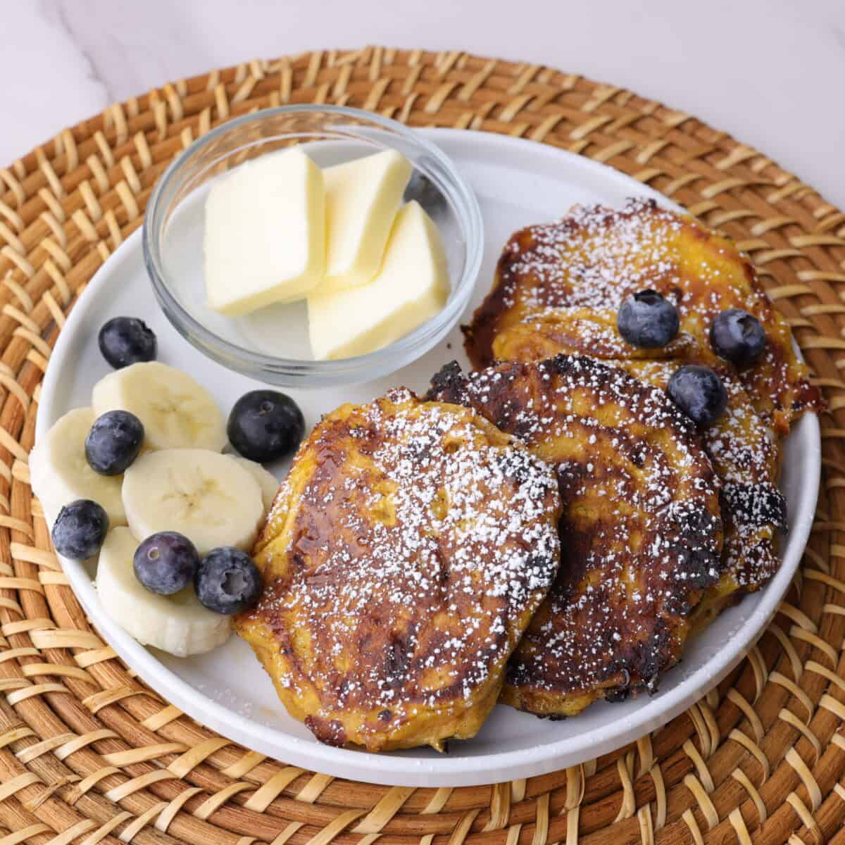 Perfectly browned pumpkin cottage cheese pancakes on a plate, sprinkled with powdered sugar, topped with blueberries and bananas and a small dish of cubed butter on the side.