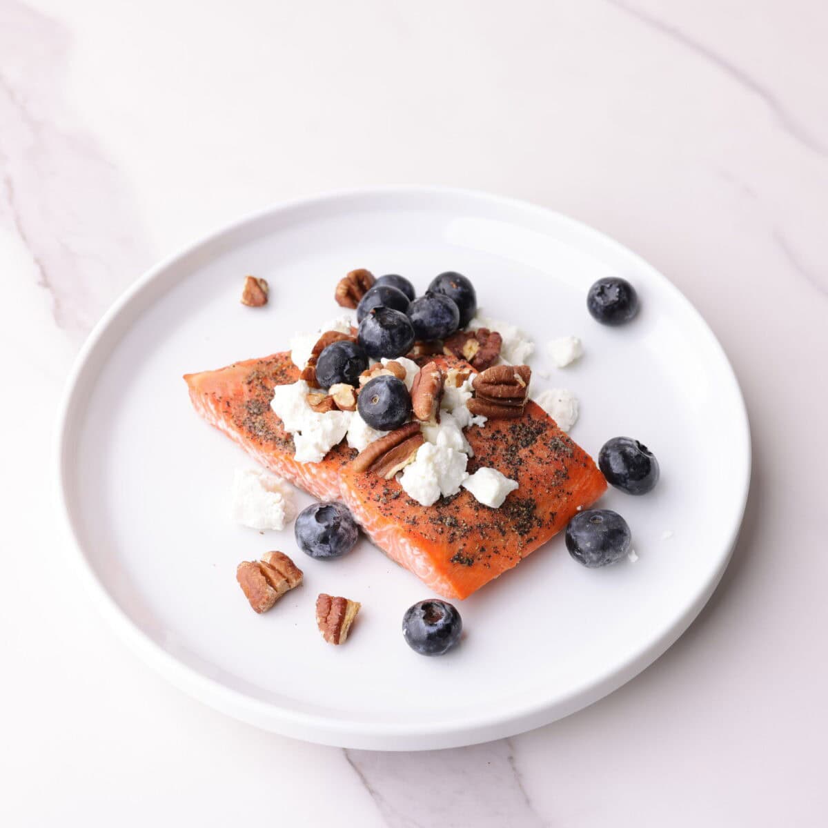 Perfectly baked salmon topped with goat cheese, pecans, and blueberries on white plate.