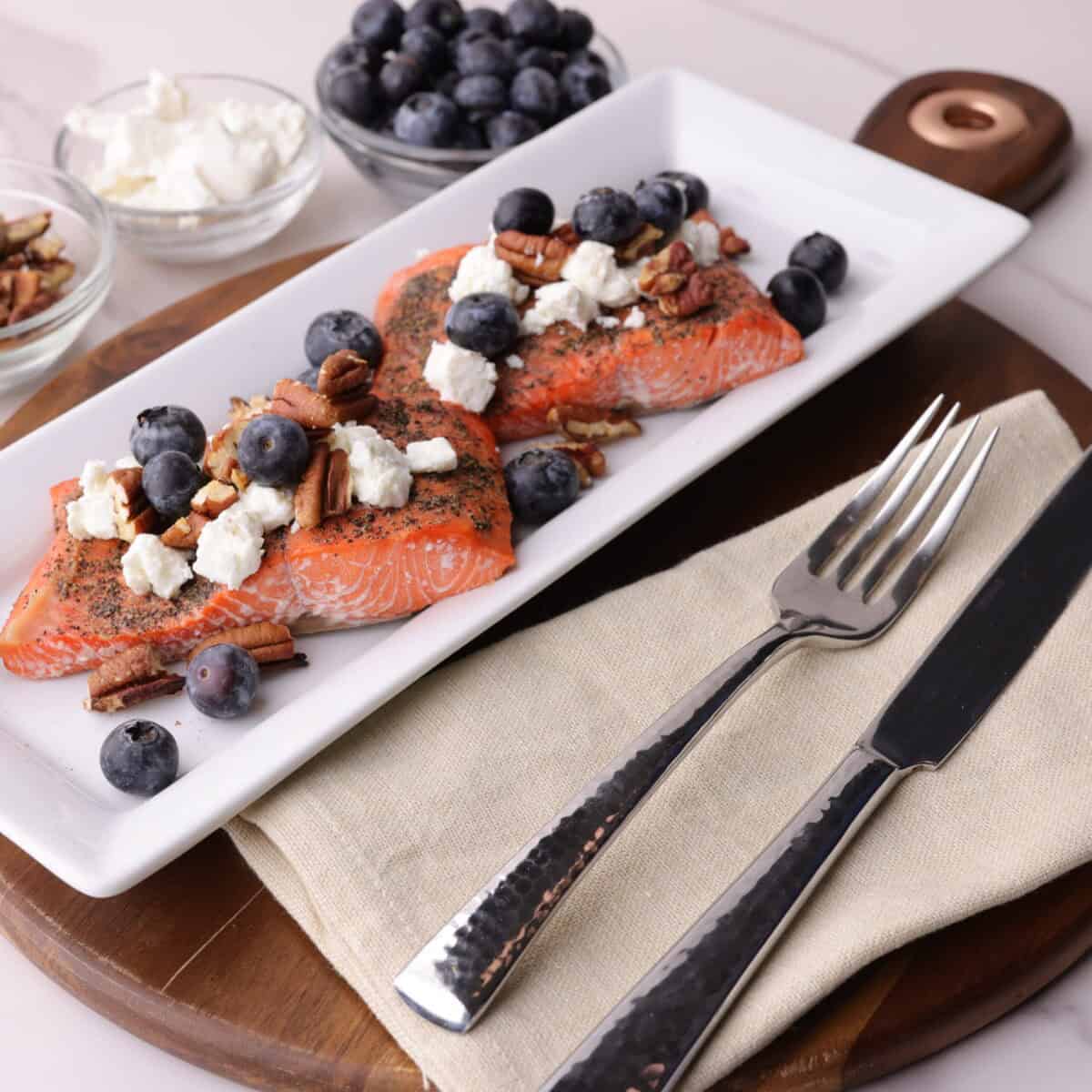 Two cooked salmon fillets on a plate and topped with goat cheese, fresh blueberries and roasted pecans. The plate is decoratively placed on a charcuterie board with a cloth napkin, fork and knife with bowls of blueberries, goat cheese and pecans in the background for decoration. 