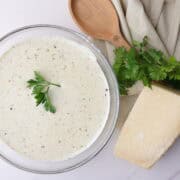 A bowl of creamy homemade Alfredo sauce with cream cheese topped with parsley and positioned decoratively next to a napkin, spoon and block of Parmesan cheese.
