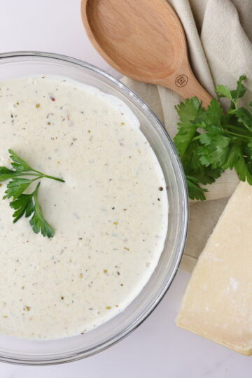 A bowl of creamy homemade Alfredo sauce with cream cheese topped with parsley and positioned decoratively next to a napkin, spoon and block of Parmesan cheese.