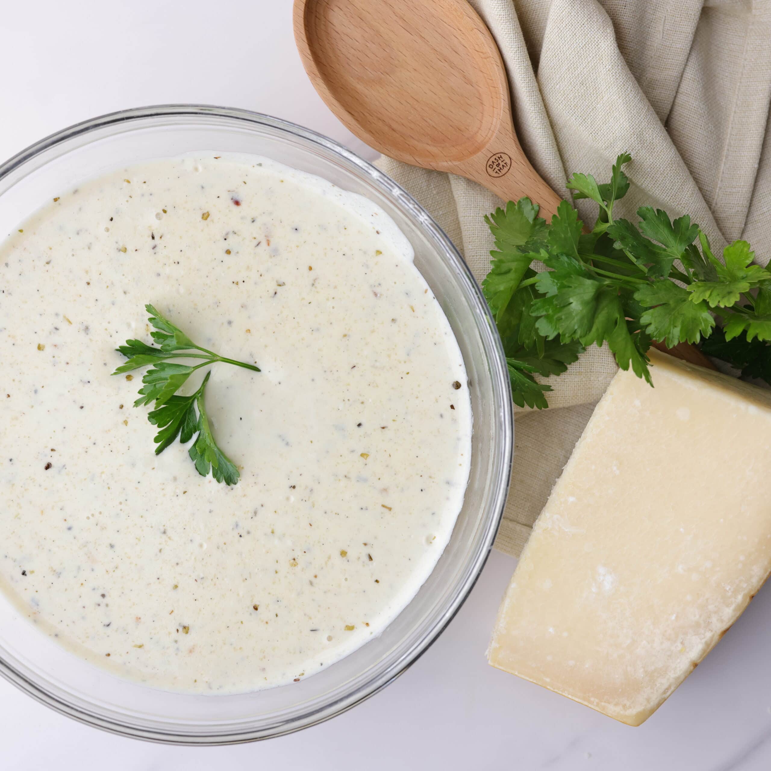 A bowl of creamy homemade Alfredo sauce with cream cheese topped with parsley and positioned decoratively next to a napkin, spoon and block of Parmesan cheese. 