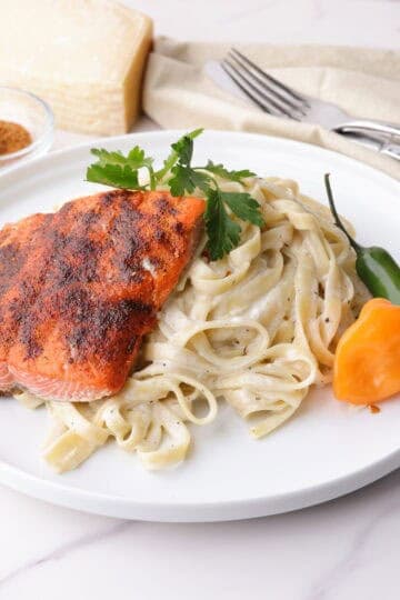 A plate of creamy fettuccine Alfredo topped with a 3 ounce salmon fillet with blackened seasoning topped with parsley and accented with a habanero and serrano pepper. In the background is a bowl of blackened seasoning and a block of Parmesan cheese.