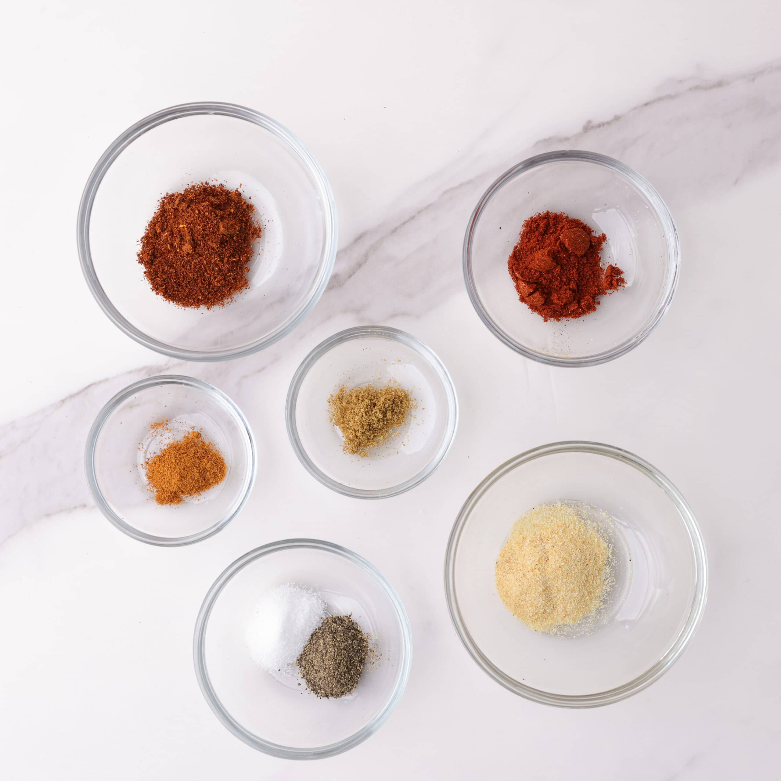 Individual glass bowls of the spices needed for the blackened seasoning - chili powder, smoked paprika, cayenne pepper, cumin, garlic powder, salt and pepper.
