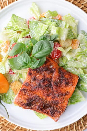 A plate of crisp romaine lettuce, mixed with sweet bell peppers, cherry tomatoes and seasoned croutons, all tossed in Caesar dressing and topped with a perfectly baked blackened salmon fillet. Garnished with basil and spicy peppers.