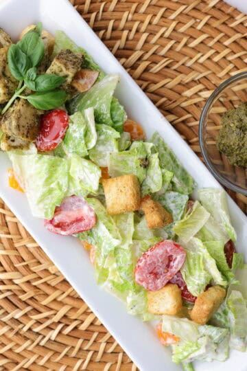 Mixed Caesar salad plated with pesto chicken on the side and topped with basil.