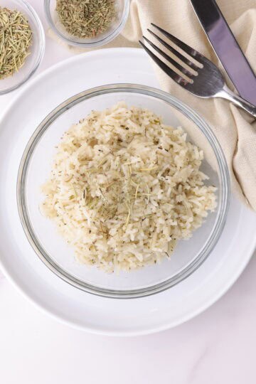 Tender basmati rice, cooked with the aromatic additions of rosemary and thyme., displayed in a bowl with a fork and knife, topped with rosemary and bowls of rosemary and thyme beside it for visual appeal.