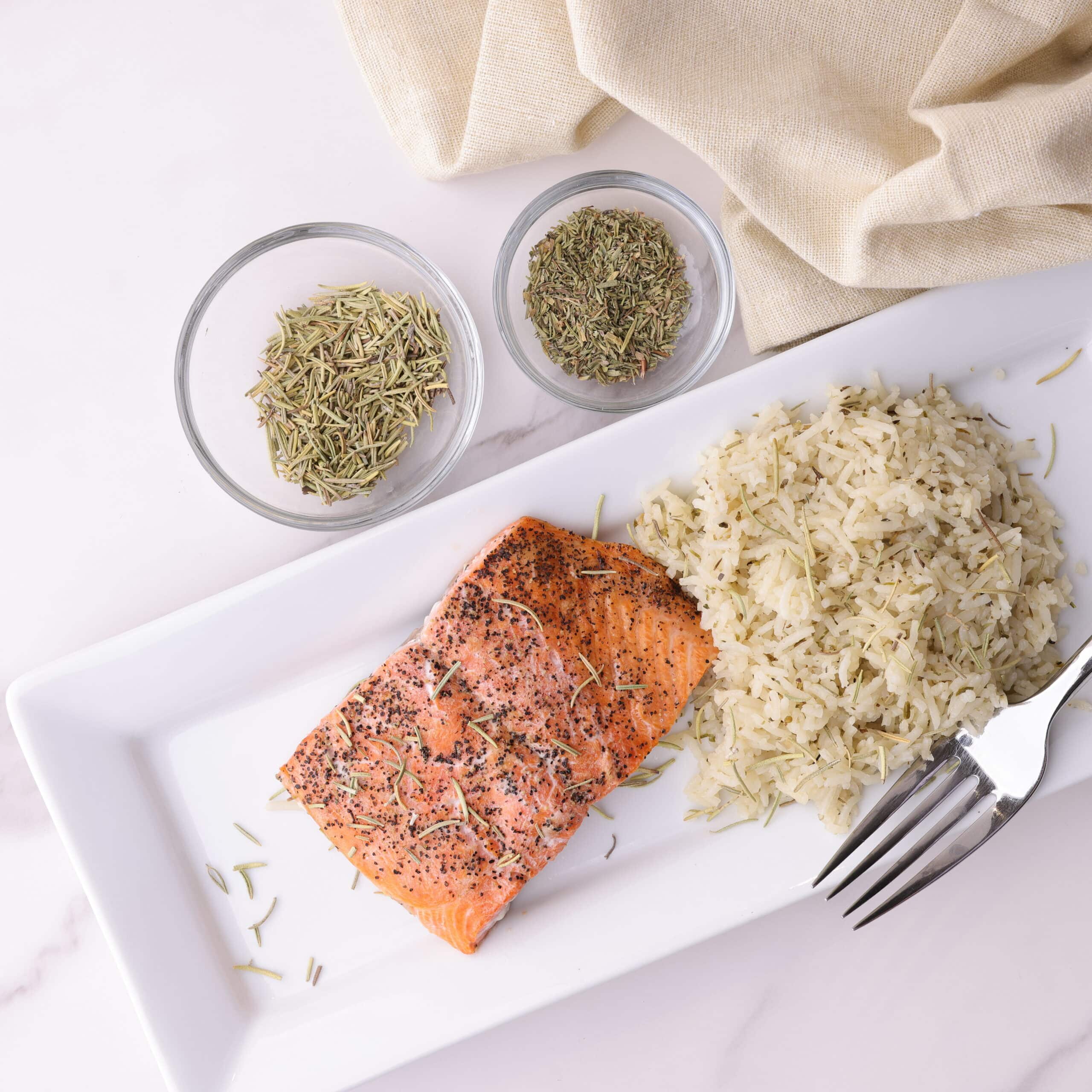 A display of the cooked rosemary basmati rice on a plate with a cooked and lightly seasoned fillet of salmon, sprinkled with rosemary and a fork at its side. 