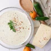 A bowl of creamy spicy Alfredo sauce topped with parsley and red pepper chili flakes, positioned decoratively next to a napkin, spoon, block of Parmesan cheese and jalapeño, habanero and serrano peppers to demonstrate it's spicy undertones.