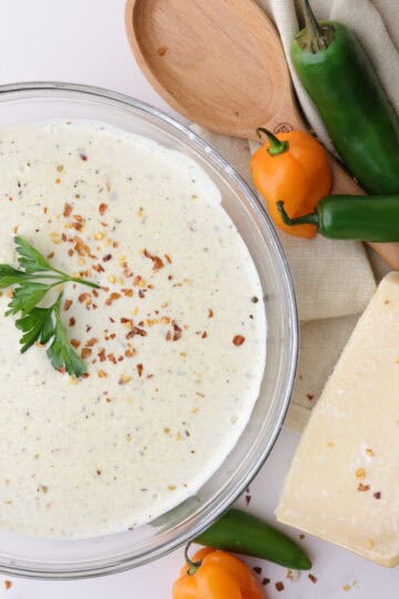 A bowl of creamy spicy Alfredo sauce topped with parsley and red pepper chili flakes, positioned decoratively next to a napkin, spoon, block of Parmesan cheese and jalapeño, habanero and serrano peppers to demonstrate it's spicy undertones.