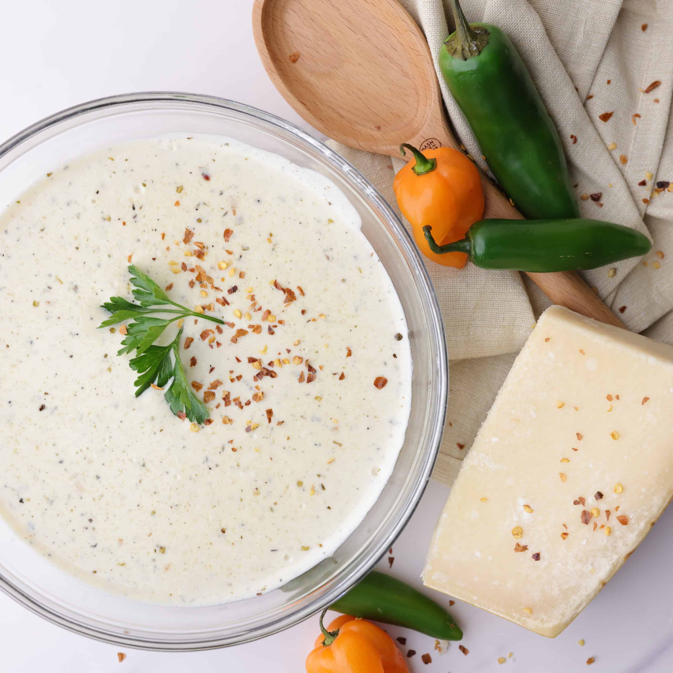 A bowl of creamy spicy Alfredo sauce topped with parsley and red pepper chili flakes, positioned decoratively next to a napkin, spoon, block of Parmesan cheese and jalapeño, habanero and serrano peppers to demonstrate it's spicy undertones.