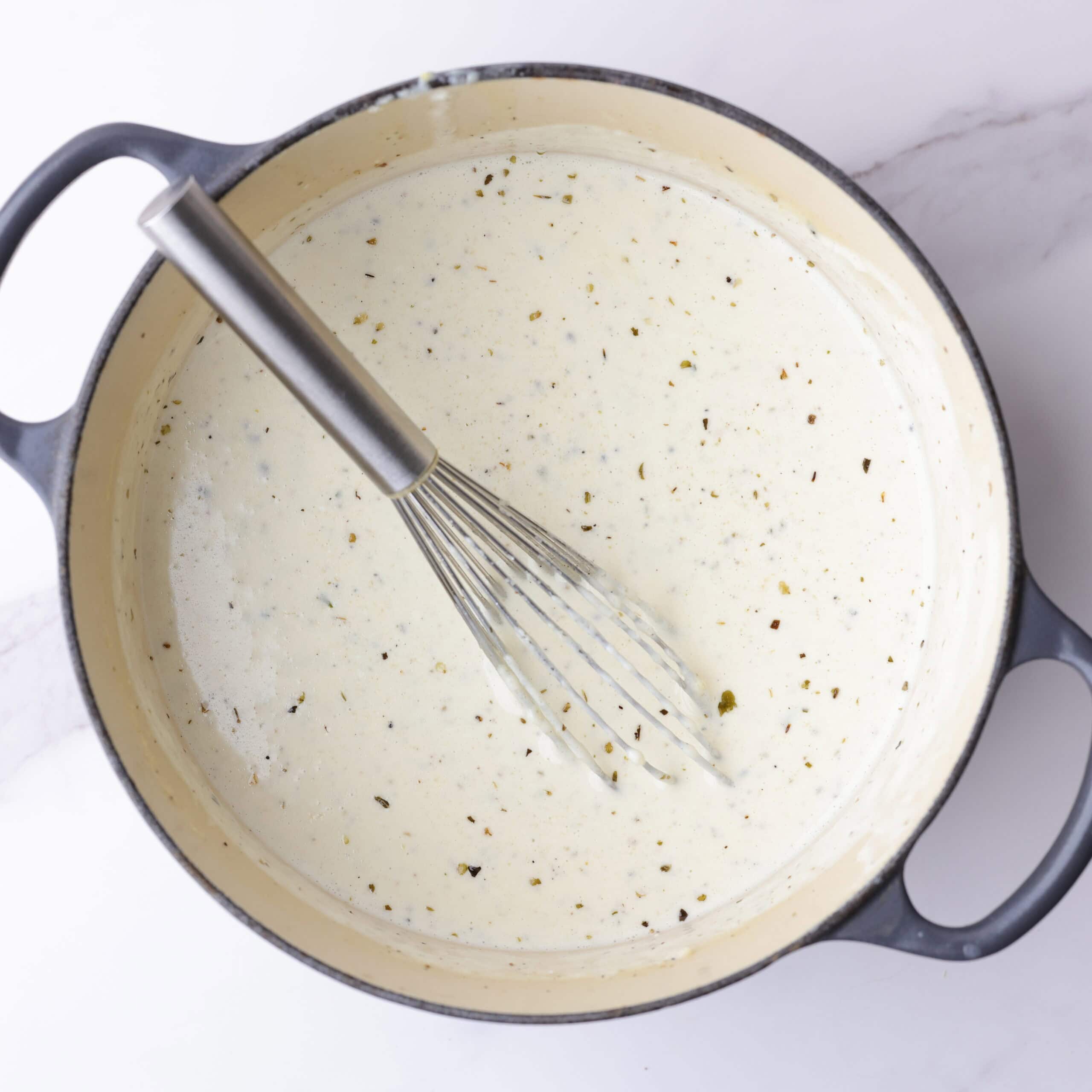 A small pot filled with thick and creamy homemade Alfredo sauce with a whisk in it.