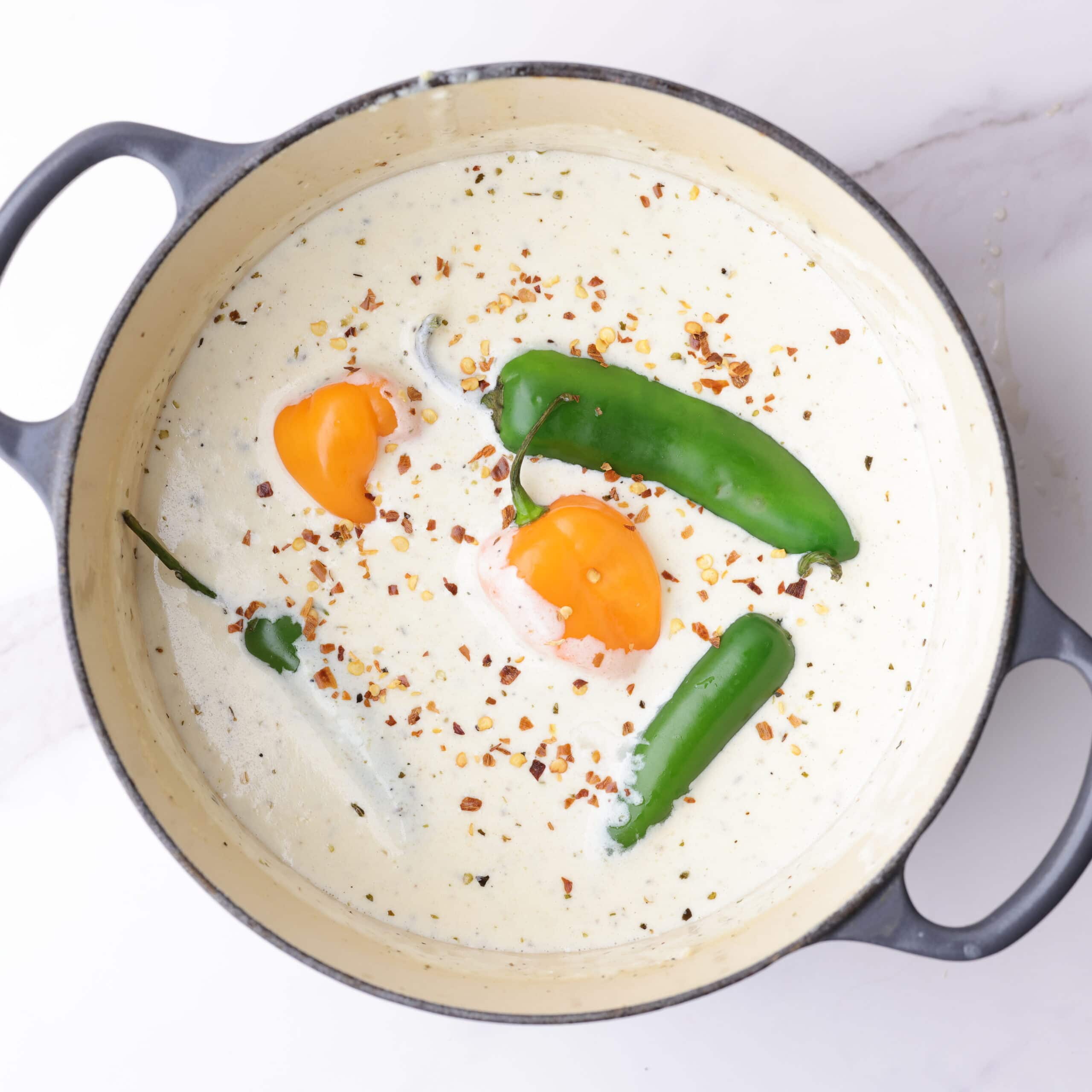 A small pot filled with thick and creamy Alfredo sauce with whole jalapeño, habanero and serrano peppers and red pepper chili flakes floating on top to be simmered into the sauce so as to imbue their heat into it.