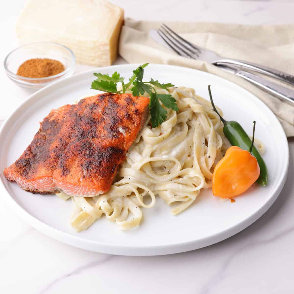 A plate of creamy fettuccine Alfredo topped with a 3 ounce salmon fillet with blackened seasoning topped with parsley and accented with a habanero and serrano pepper. In the background is a bowl of blackened seasoning and a block of Parmesan cheese.