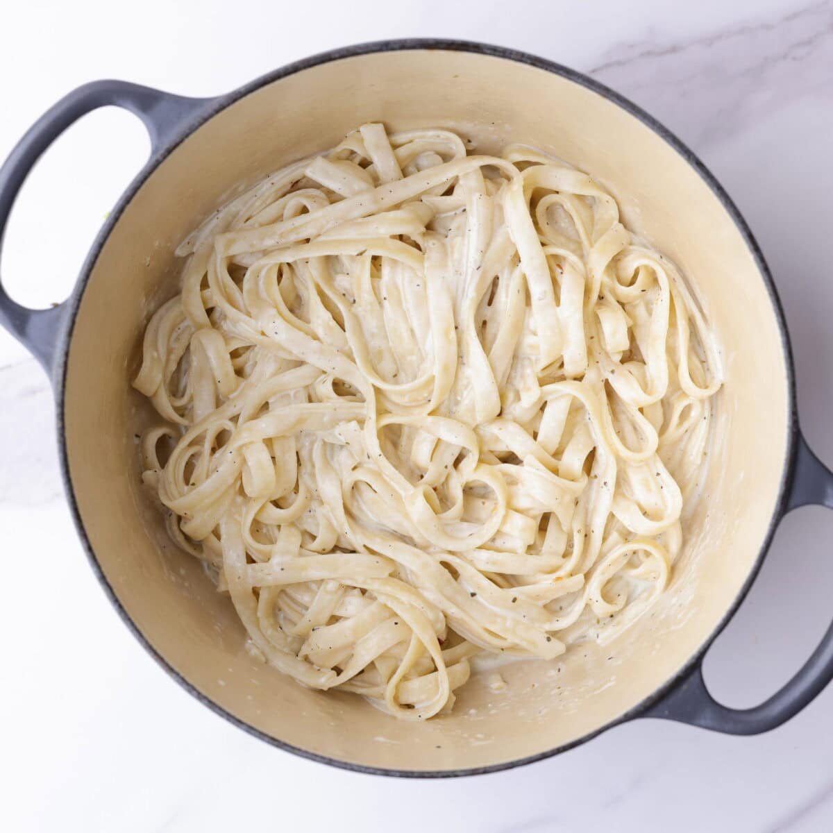 A small enameled pot with tenderly cooked fettuccine noodles covered in creamy Alfredo sauce.