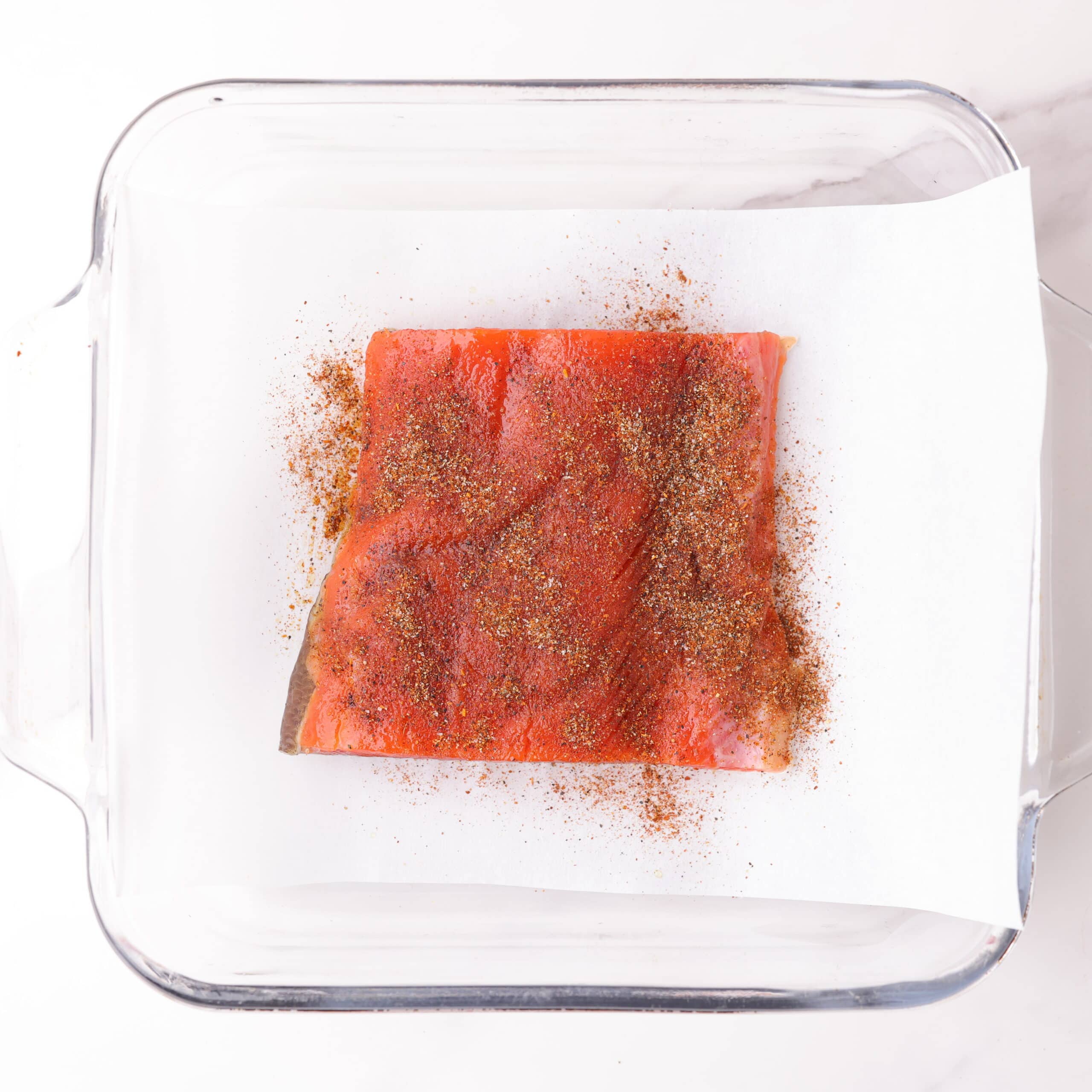 A 4 ounce red salmon fillet in a glass baking dish on parchment paper, lightly oiled and sprinkled with the blackened seasoning mix.