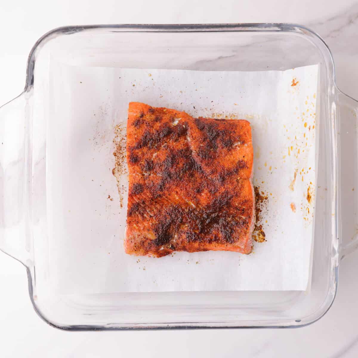 A blackened seasoned 4 ounce red salmon fillet in a glass baking dish on parchment paper, baked to light flakiness. 
