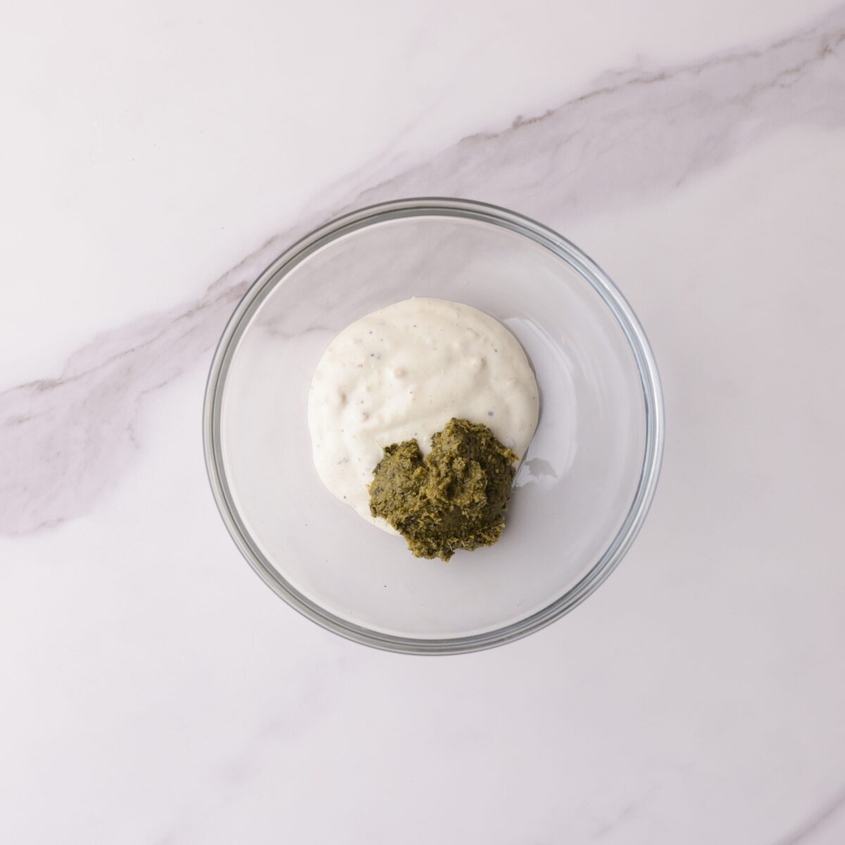A small glass bowl with Caesar salad dressing and basil pesto side by side, ready to be mixed together.
