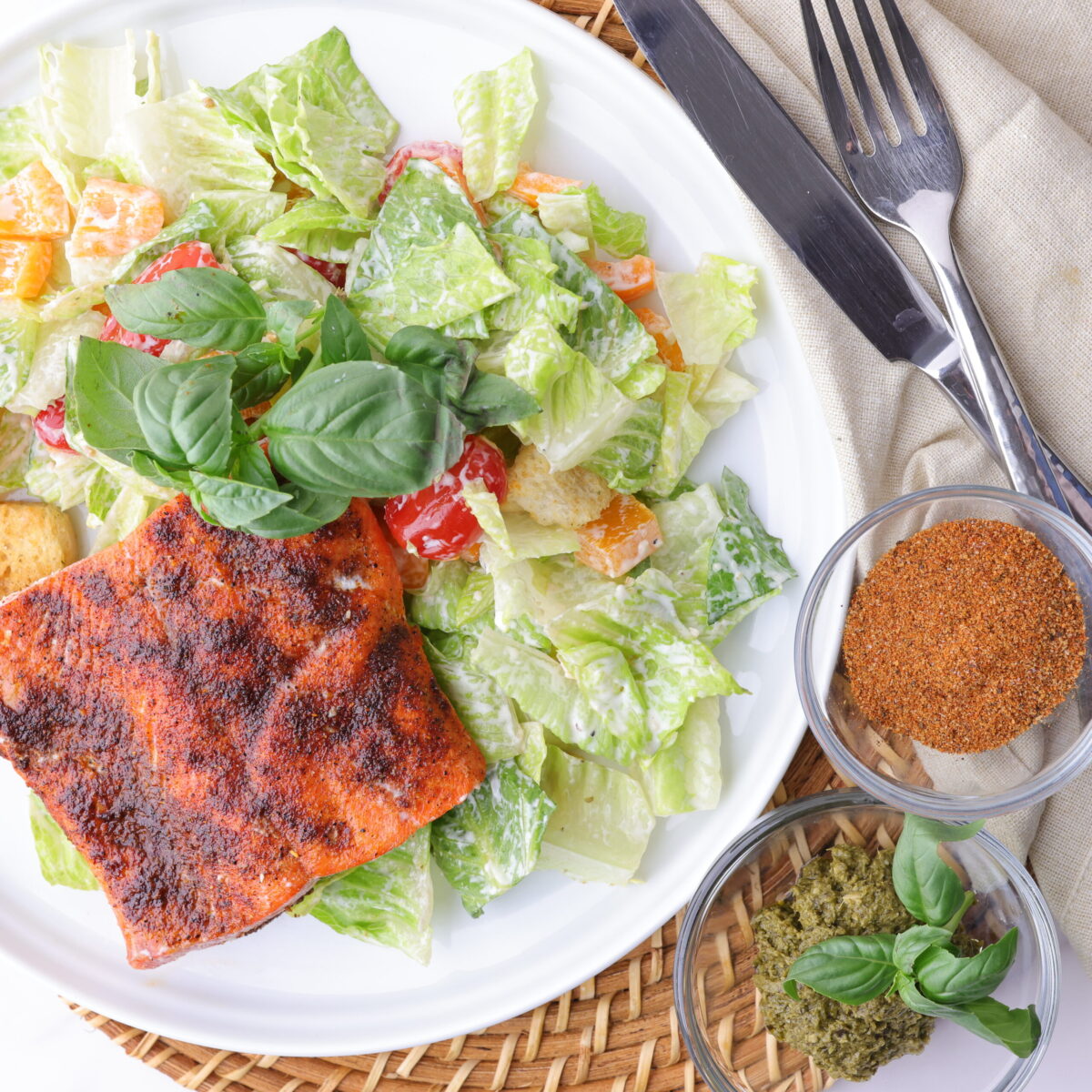 A plate of crisp romaine lettuce, mixed with sweet bell peppers, cherry tomatoes and seasoned croutons, all tossed in Caesar dressing and topped with a perfectly baked blackened salmon fillet. Garnished with basil and set with cutlery, a napkin and bowls of seasoning and pesto.