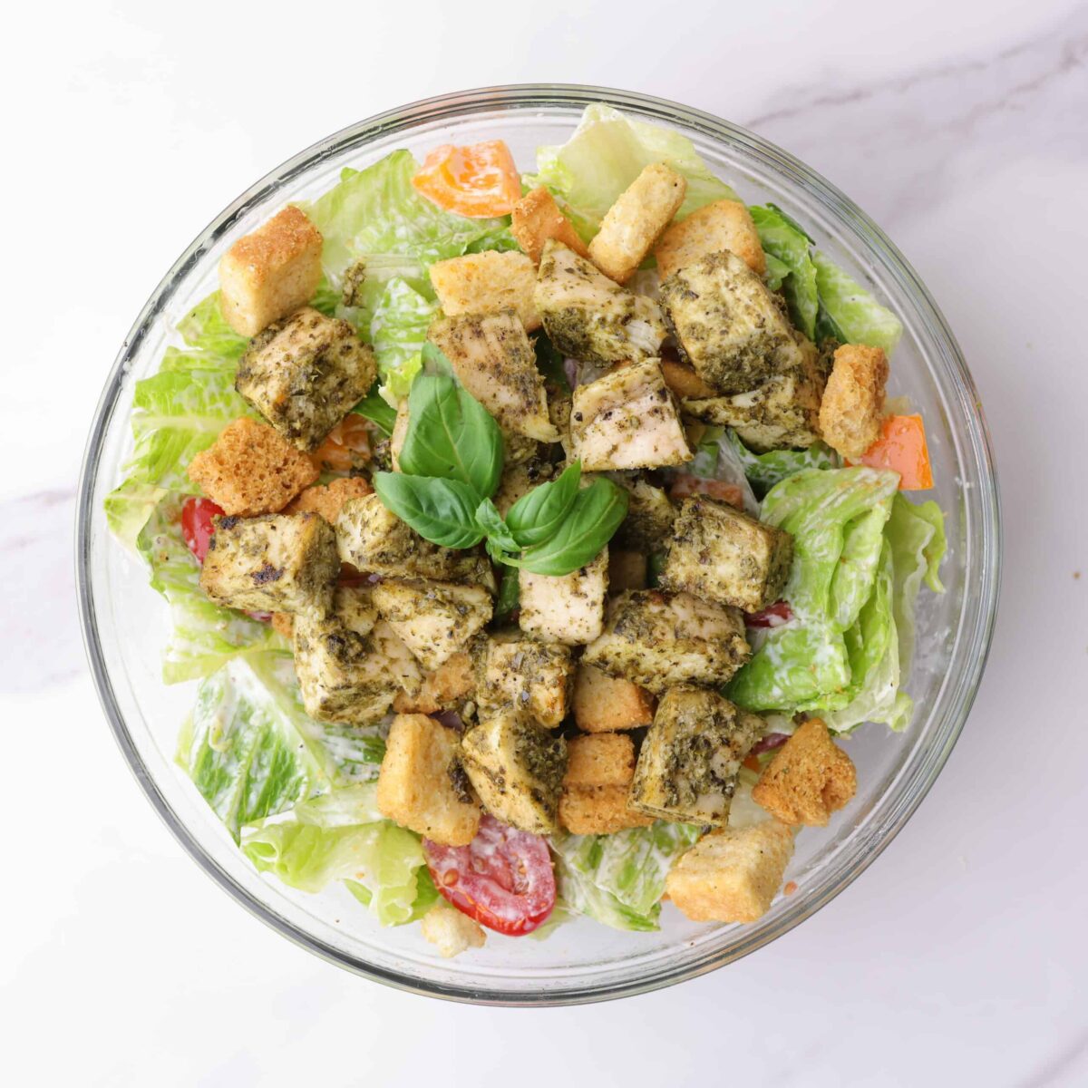 A completed bowl of salad tossed with dressing and veggies, topped with croutons and pesto chicken and garnished with basil.