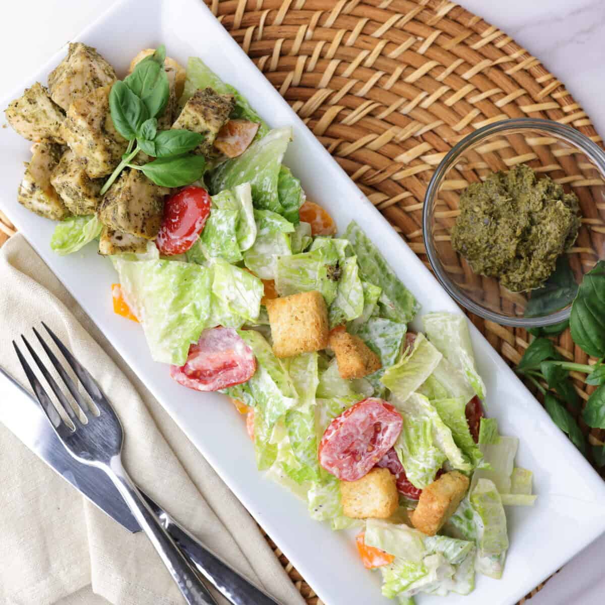 Mixed Caesar salad plated with pesto chicken on the side and topped with basil.