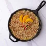 Skillet with golden brown rhubarb peach crisp topped with sliced peaches and rhubarb.