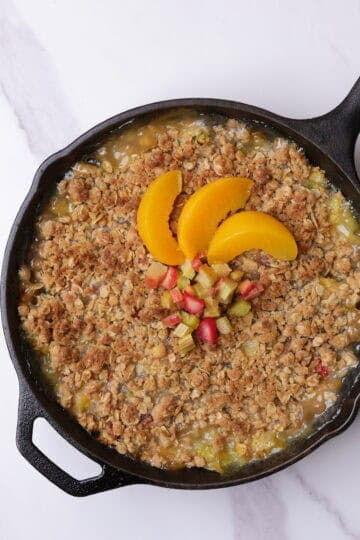 Skillet with golden brown rhubarb peach crisp topped with sliced peaches and rhubarb.